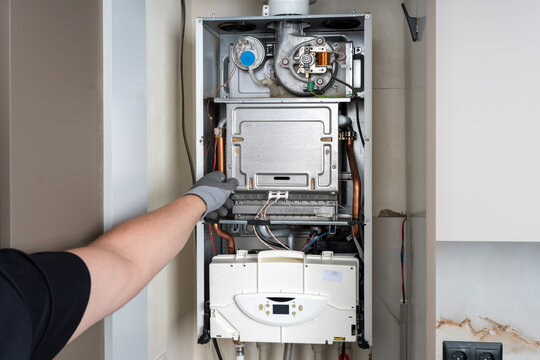 Technician in a protective glove pointing at internal components and mechanisms of an open domestic gas boiler during maintenance or repair service.