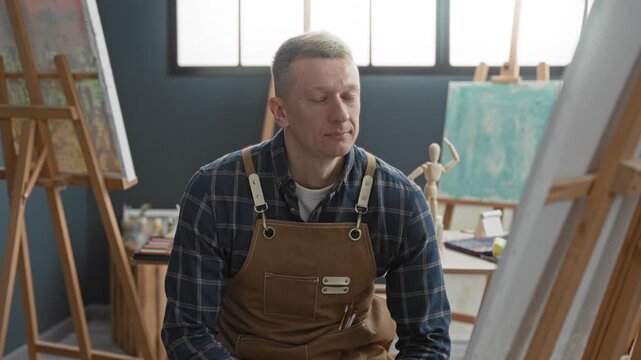 Man painter in apron smiles seated by wooden easel, hand holding brush and palette, surrounded by canvases and art supplies in studio; creative contentment.