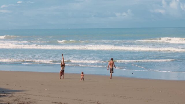 Family of three plays on wide sandy tropical beach with gentle ocean waves rolling in, woman performs cartwheels while father watches toddler near the water under blue sky with scattered clouds.