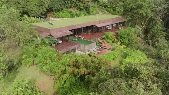 Girl swims in pool of luxurious villa nestled in lush tropical jungle. Aerial drone view reveals modern architecture with green roof and wooden deck surrounded by dense rainforest vegetation.