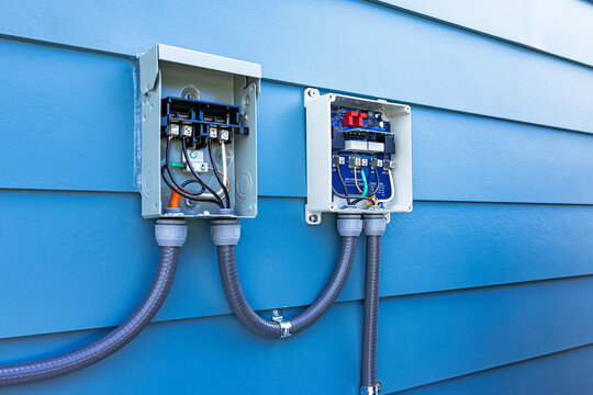 Perspective view of an open AC disconnect box and surge protector wired with flexible conduit on a blue siding house wall outdoors