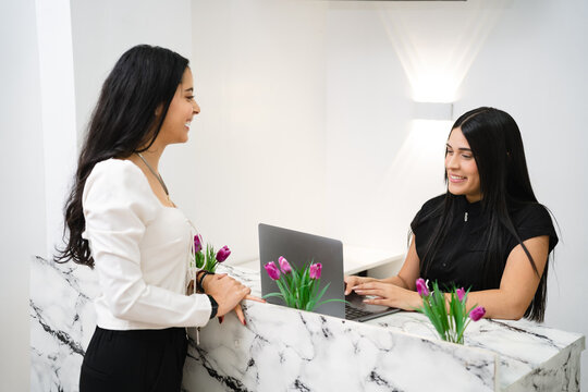 Two young adult Hispanic women communicating at a modern marble reception desk with a laptop.