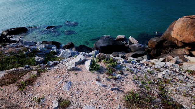 People walk near the rocks by the ocean in Dak Lak. The blue water stretches far into the distance. The sky is clear and bright. The calming waves gently hit the shore.