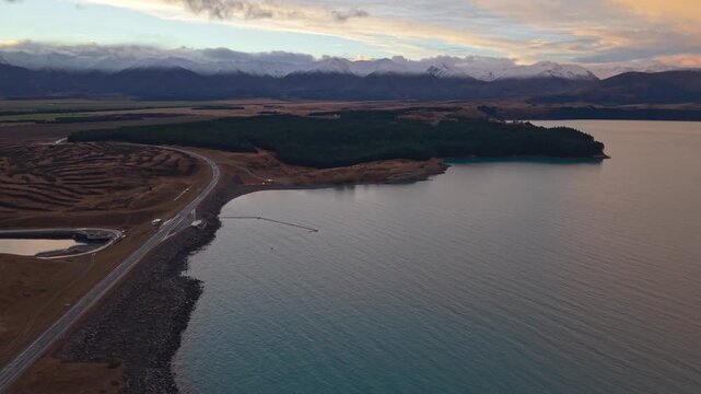 Aerial view of Lake Tekapo shoreline, road, and distant snowy mountains at sunset