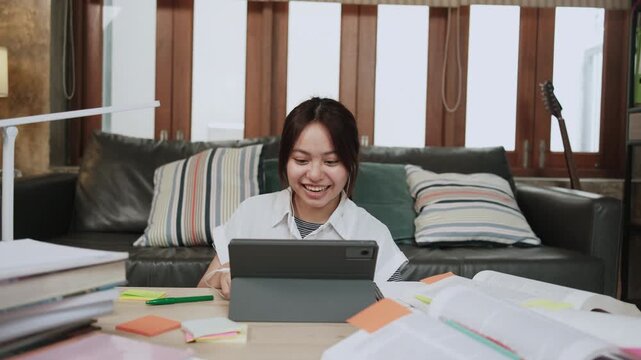Asian teenage girl greeting in online class, remote learning at home using digital tablet, writing and reading textbooks, self-study for back to school, Homeschooling, university education lifestyle.