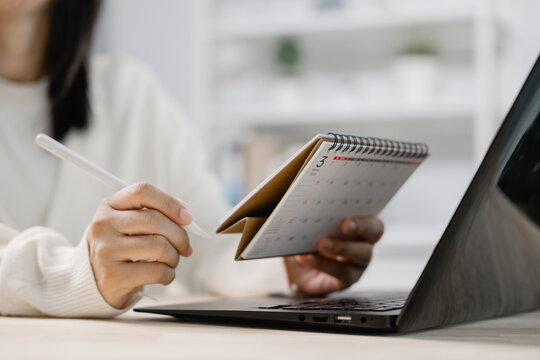 Hand with pen on calendar paper at office desk planning schedule date, while woman holds monthly organizer near open laptop computer to manage important work appointments