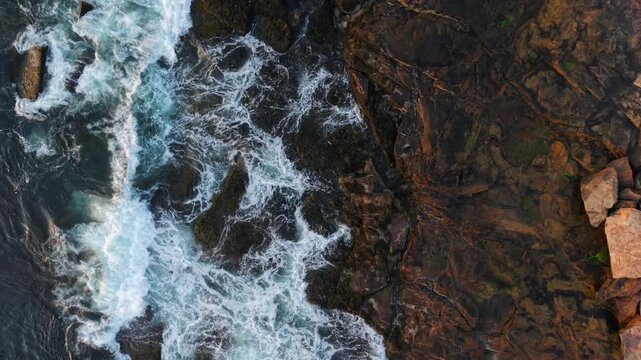 Top-down drone fly over Atlantic Ocean water surf crashes against a dark rocky Gloucester shoreline, Massachusetts, US