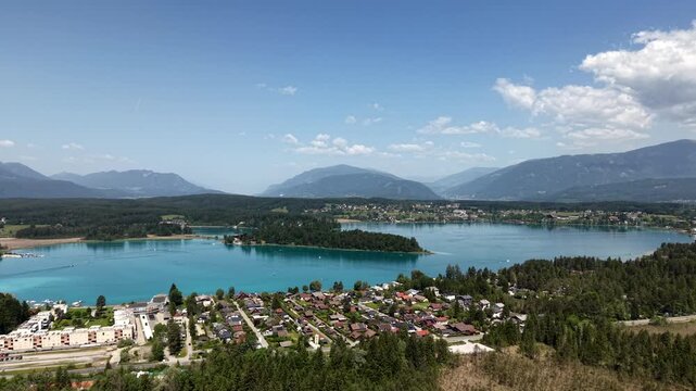 Turquoise Faaker See stretches beneath towering Austrian Alps, lakeside neighborhood houses nestled among dense pine forest near small wooded island in calm summer waters. Aerial drone wide shot