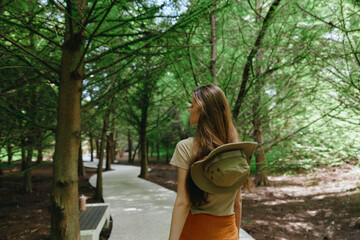 Naklejka premium Woman walking on forest trail with backpack and hat, back view on shaded park path surrounded by green trees and nature, dappled sunlight and casual summer outdoor stroll.