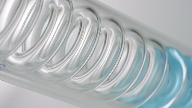 Extreme close-up shot of pale blue transparent liquid flowing through the spiral of an inclined coil laboratory condenser, filling it up on grey background | Hyaluronic acid cosmetic material concept