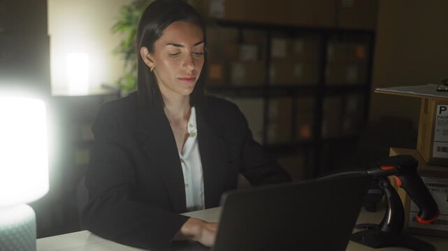 Woman at laptop typing then stretching arms overhead amid shelving and boxes in warehouse building; late night relief.