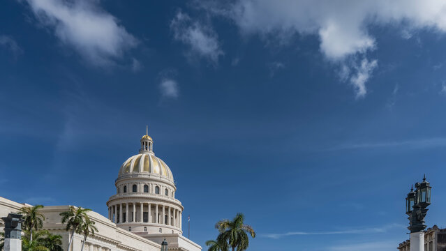 The Capitol building in Havana. A gilded dome with a spire, colonnade, arches against a background of blue sky and clouds. Palm trees. Cuba.