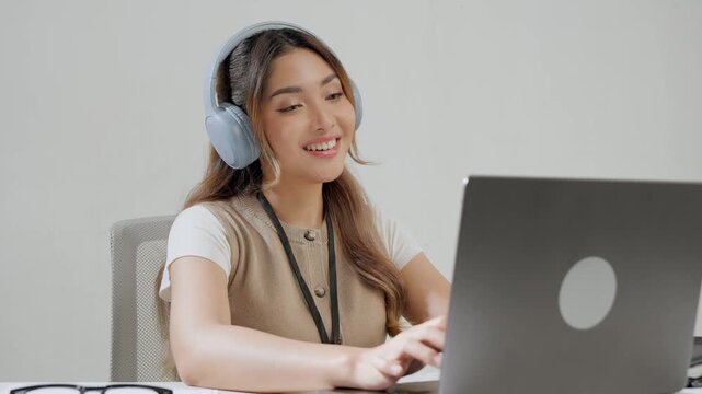 Happy young Asian woman is wearing headphone listening to music while working using her laptop.