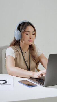 Happy young Asian woman is wearing headphone listening to music while working using her laptop.