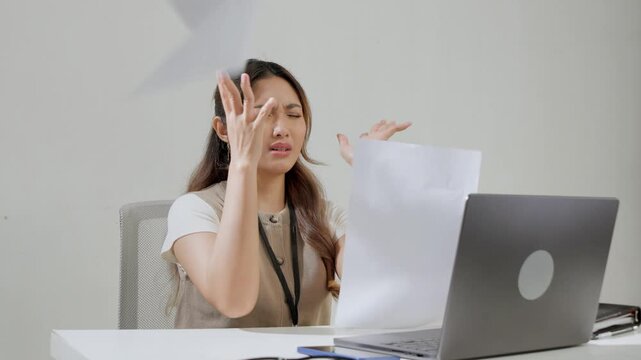 Stressful young Asian woman is sitting while throwing paperworks with frustrated expression.