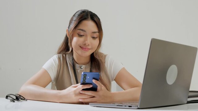 Cheerful young Asian woman is working while using smartphone to text someone or open social media.