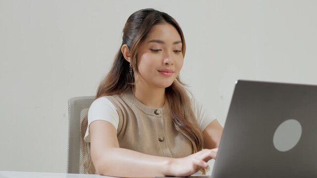 Focused young Asian woman is typing report on her laptop while feeling happy.