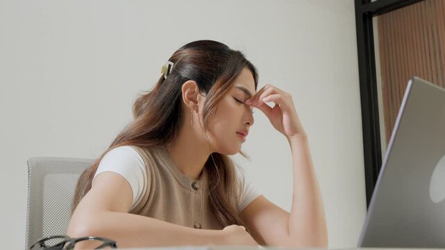 Young Asian female employee is working using her laptop while feeling headache and stressful.
