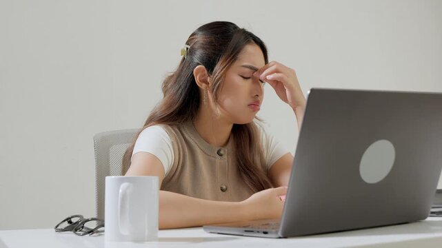 Young Asian female employee is working using her laptop while feeling headache and stressful.