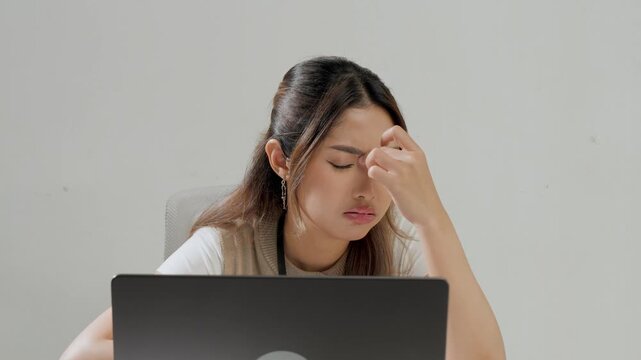Young Asian female employee is working using her laptop while feeling headache and stressful.