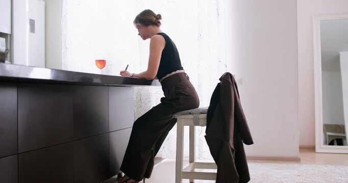 Quiet routine. Daily planning. Thoughtful stylish woman writing notes at home table enjoying orange drink in glass interior.