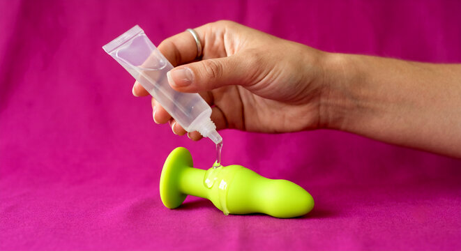 A hand applies clear lubricant from a small squeeze tube onto a bright lime green silicone butt plug, all set against a vibrant magenta pink background.