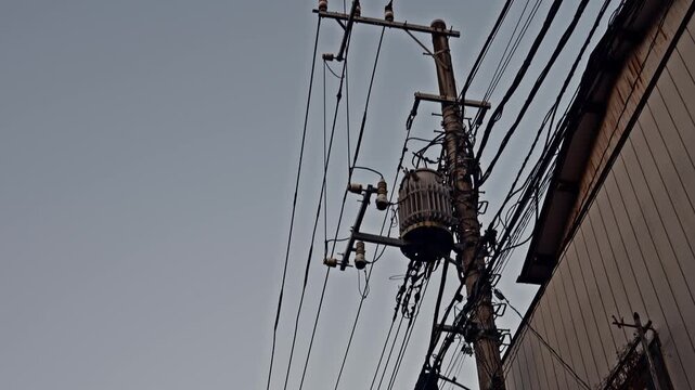 A weathered concrete pole bearing a large cylindrical transformer and tangled cable connections fills a narrow gap between buildings beneath a clear gray dusk sky.