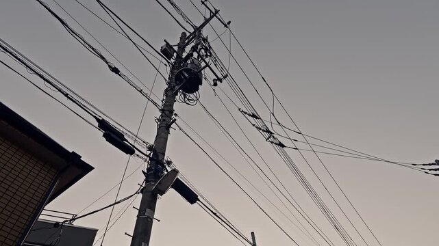 A concrete pole bearing a transformer and branching cable connections rises beside a brick building corner. Multiple wires radiate outward against a pale gray dusk sky.