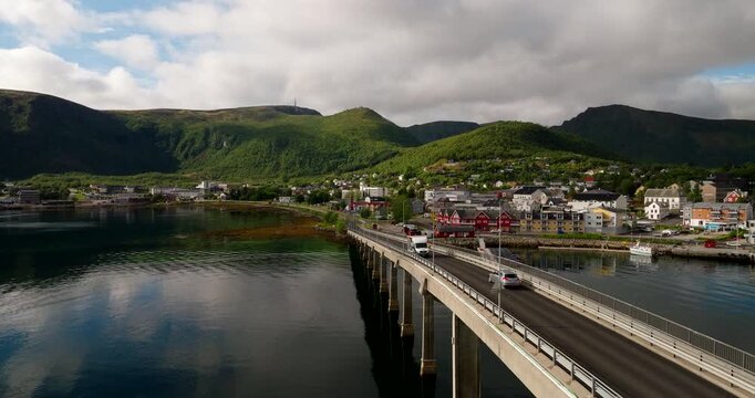 Vehicle crossing a coastal bridge toward a waterfront town surrounded by mountains. Captured in Stokmarknes, Norway, showcasing transport, travel, and scenic infrastructure, aerial dolly view