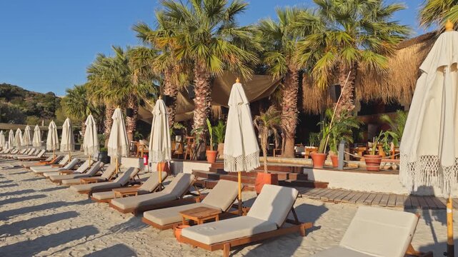 Static wide shot of rows of empty wooden sunbeds and closed boho fringe umbrellas on a sandy beach at a luxury coastal resort during peaceful morning golden hour.