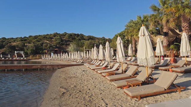 Wide shot of a quiet beach club at golden hour, rows of sun loungers and folded umbrellas along a sandy shore, wooden pier extending into a calm bay.