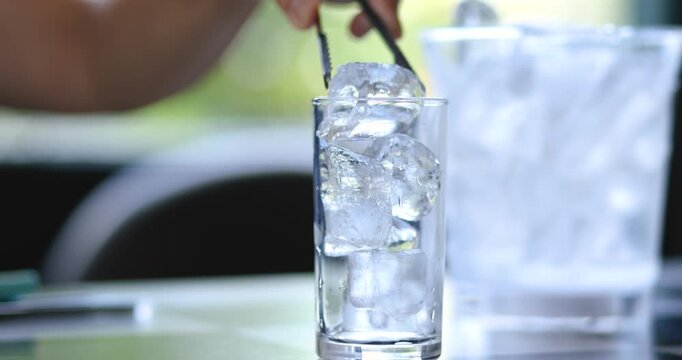 Hand using tongs to place ice cubes into a glass from an ice bucket. Beverage preparation, Bar service, Refreshment, Hospitality and food service concepts, close-up
