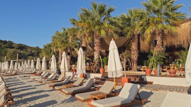 Static wide shot of rows of empty wooden sunbeds and closed boho fringe umbrellas on a sandy beach at a luxury coastal resort during peaceful morning golden hour.