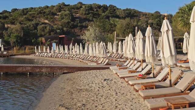 Wide shot of a quiet beach club at golden hour, rows of sun loungers and folded umbrellas along a sandy shore, wooden pier extending into a calm bay.