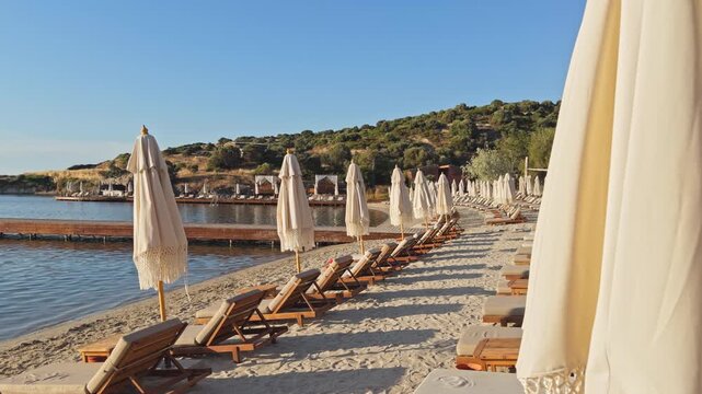 Static wide shot of rows of empty wooden sunbeds and closed boho fringe umbrellas on a sandy beach at a luxury coastal resort during peaceful morning golden hour.