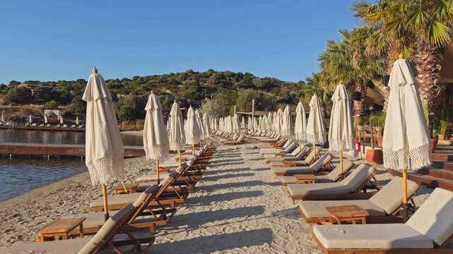 Static wide shot of rows of empty wooden sunbeds and closed boho fringe umbrellas on a sandy beach at a luxury coastal resort during peaceful morning golden hour.