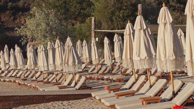 Static wide shot of rows of empty wooden sunbeds and closed boho fringe umbrellas on a sandy beach at a luxury coastal resort during peaceful morning golden hour.