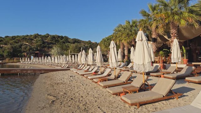 Wide shot of a quiet beach club at golden hour, rows of sun loungers and folded umbrellas along a sandy shore, wooden pier extending into a calm bay.
