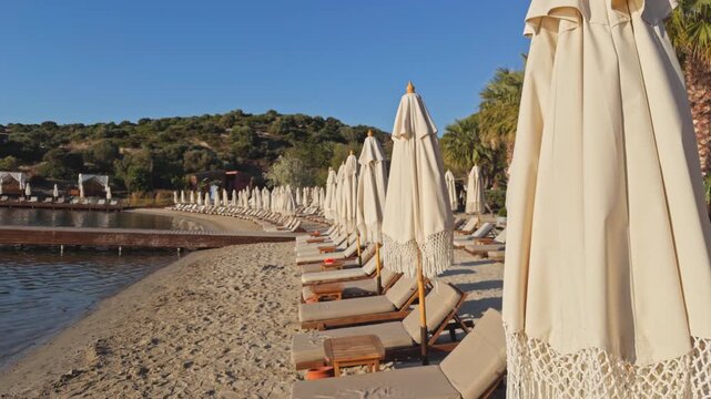 Wide shot of a quiet beach club at golden hour, rows of sun loungers and folded umbrellas along a sandy shore, wooden pier extending into a calm bay.