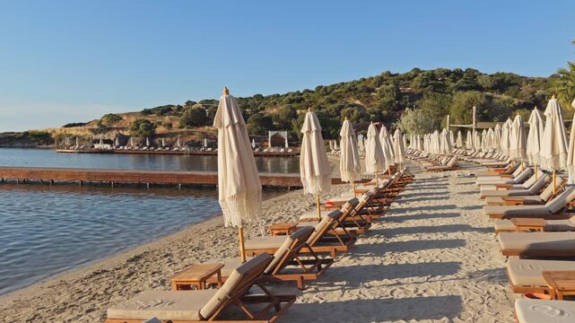 Static wide shot of rows of empty wooden sunbeds and closed boho fringe umbrellas on a sandy beach at a luxury coastal resort during peaceful morning golden hour.