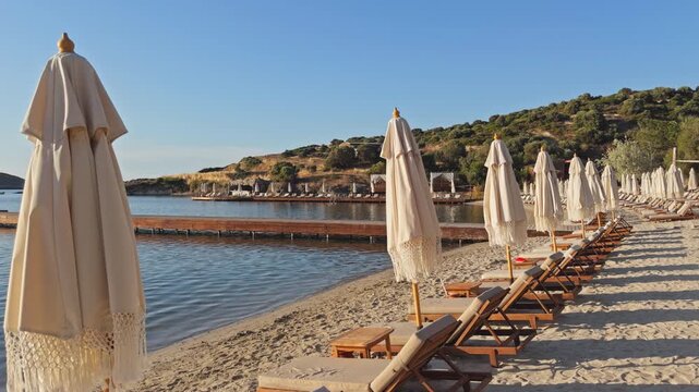 Static wide shot of rows of empty wooden sunbeds and closed boho fringe umbrellas on a sandy beach at a luxury coastal resort during peaceful morning golden hour.