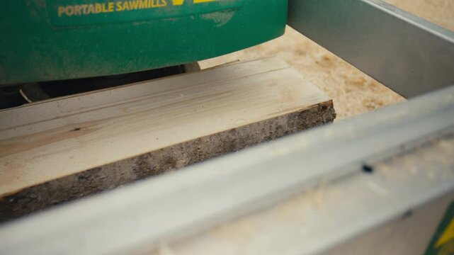 A slow motion detail shot shows a freshly cut board emerging from a portable sawmill in an outdoor setting, emphasizing pale grain, rough bark edge and drifting sawdust.