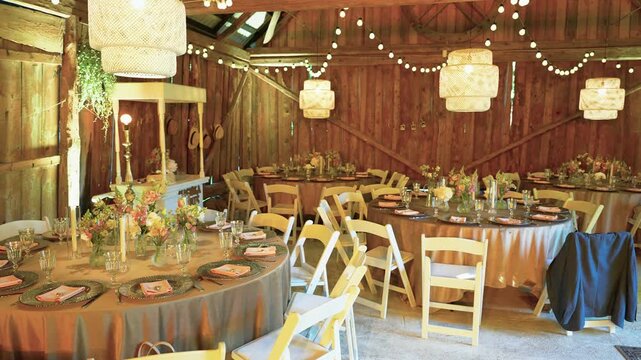Charming rustic barn wedding reception features round tables with floral centerpieces, elegant place settings, string lights, and lanterns. Warm ambient lighting creates a cozy, festive atmosphere.