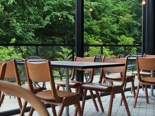 Obraz premium Empty chairs line a modern cafe patio. Large windows frame lush green forest beyond. Natural light bathes the scene in calm tranquility. Wooden tables and chairs invite quiet conversation