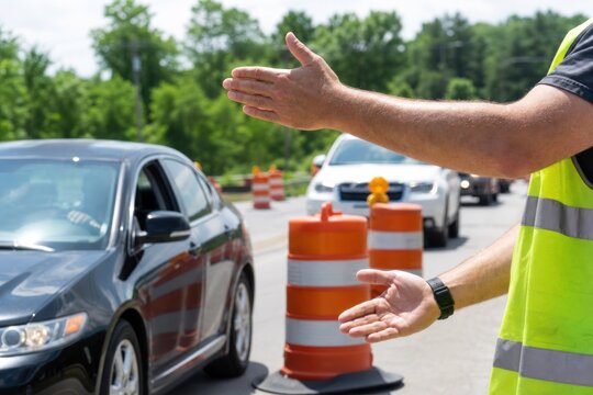Construction worker in high-visibility vest directing traffic flow on road with orange safety barrels, professional flagger managing vehicle movement at highway work zone, safety and infrastructure.