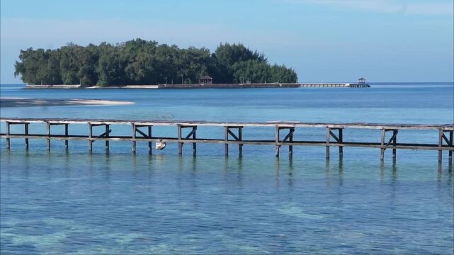 Static scenic footage of a wooden jetty extending across crystal-clear turquoise water toward a lush tropical island under a clear blue sky. Peaceful coastal landscape ideal for travel and tourism.