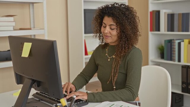 Woman waving hand and typing at computer in home office, smiling toward camera at tidy desk with books and papers; friendly welcome.
