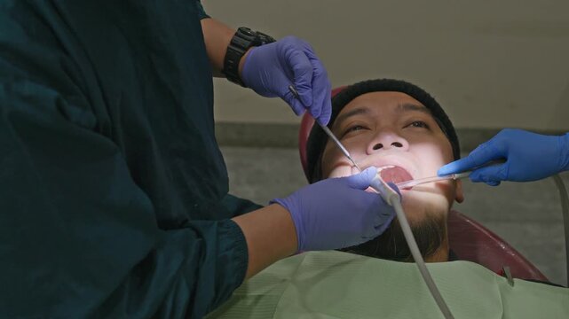 Dentist and assistant using saliva ejector and mirror during male patient's teeth checkup