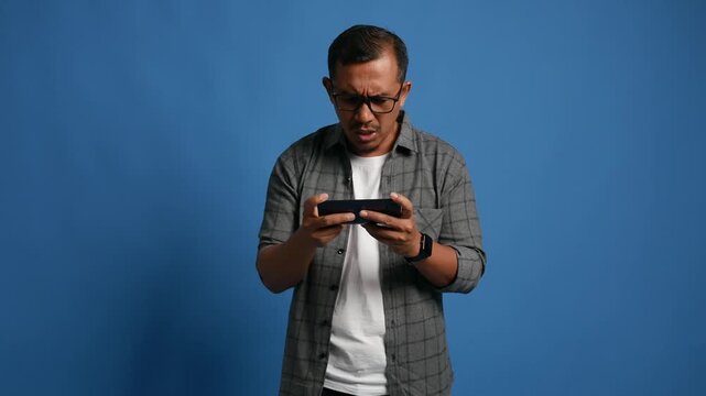 An adult man standing isolated on a blue background, playing an online game on his mobile phone, with an angry expression, shouting because he lost the game.