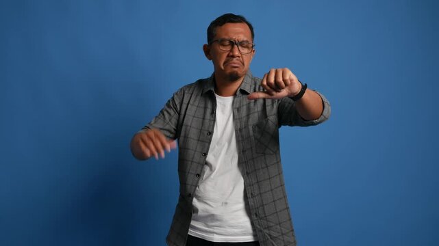 Asian adult man standing isolated on blue background, showing thumbs down with expression of displeasure, negative emotion, rejection or disagreement